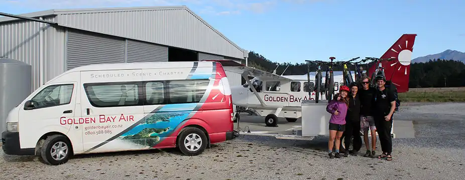 A group of three bikers pose in front of Golden Bay Air passenger van with trailer carrying mountain bikes at Takaka Airport with Airvan aircraft parked in background