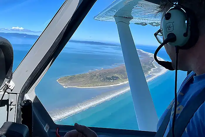 Passenger wearing a headset looking out window of airplane to Farewell Spit lighthouse