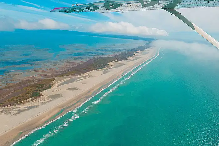 Airvan wing hovers above an expansive view of Farewell Spit bordered by tidal shallows on Golden Bay side and turquoise open sea on the outer side